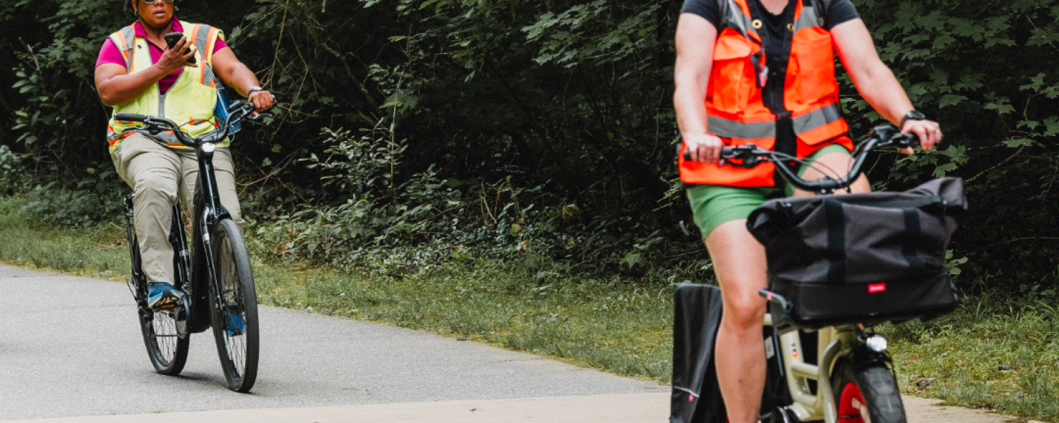 Two men in reflector vests ride bikes along a greenway path 